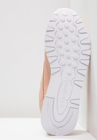 Sulan av en ljusrosa Reebok-sneaker som visar vit sula med cirkulära grepp och Reebok-logotyp, placerad vertikalt mot en vit yta.