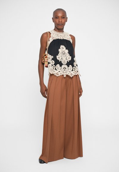 Top preto sem mangas com detalhes em renda creme, combinado com calças largas castanhas. Modelo está em pé com confiança, exibindo padrões e texturas intrincadas.