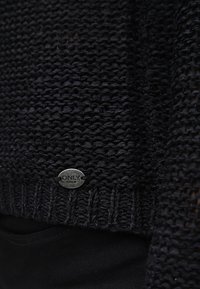 Maglione lavorato a maglia di un blu scuro con un motivo testurizzato e un'etichetta in metallo. L'orlo inferiore è a costine per un ulteriore dettaglio.