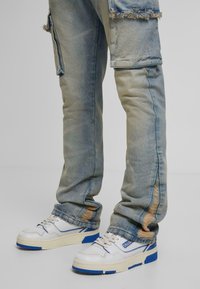 Jeans en denim bleu clair usés avec des déchirures près des genoux et des ourlets, portés avec des baskets blanches avec des accents bleus et des semelles beiges.