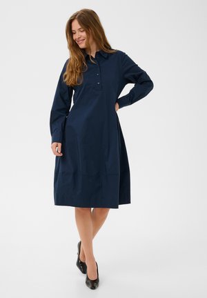 Femme aux longs cheveux bruns portant une robe chemise bleu marine à manches longues et des talons noirs, debout avec une main sur la hanche.