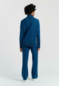 Maglione a collo alto lavorato a maglia blu e pantaloni ampi abbinati. Texture morbida, vestibilità rilassata e dettaglio con spacchi laterali sul maglione. Sneakers bianche.