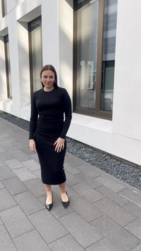 Musta pitkähihaista mekkoa, jossa on tiukka siluetti, polveen ulottuva helma, yhdistetty mustiin teräväkärkisiin korkokenkiin, taustalla modernia arkkitehtuuria.