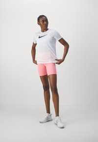 Balta īsām piedurknēm t-kreklu ar melnu Nike svilpes logotipu, saskaņots ar rozā sporta šortiem; modelis valkā baltas kurpes.