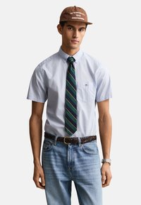 Jeune homme portant une casquette marron, une chemise bleu clair à manches courtes avec boutons, une cravate rayée, une ceinture tressée, un jean bleu clair et une montre-bracelet.