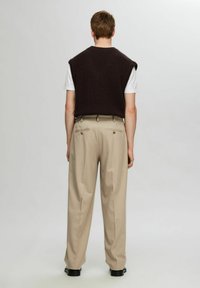 Dark brown sleeveless knit vest over a white t-shirt, paired with beige high-waisted trousers. Smooth texture, relaxed fit, and pleated design.