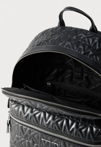 Μαύρο καπιτονέ σακίδιο πλάτης Michael Kors με ανάγλυφο μοτίβο MK, ασημένιο φερμουάρ που είναι μερικώς ανοιχτό δείχνοντας εσωτερική μαύρη επένδυση και εσωτερική τσέπη με φερμουάρ.