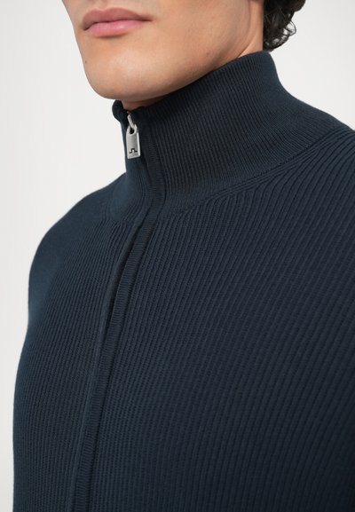 Μπλε ναυτικό ριμπ πλεκτό με ψηλό γιακά και κλείσιμο με μισό φερμουάρ. Διαθέτει υφή στο ύφασμα και λεπτομέρεια ασημένιας τράπεζας στο φερμουάρ.