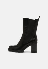 Botte noire en cuir mi-mollet avec panneaux latéraux en élastique, tirant à l'arrière, semelle crantée épaisse et talon carré sur un fond clair et uni.