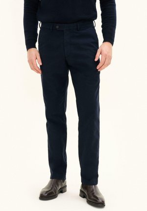 Man draagt donkerblauwe broek, zwarte laarzen en een donker lange mouwen trui, staand tegen een witte achtergrond.