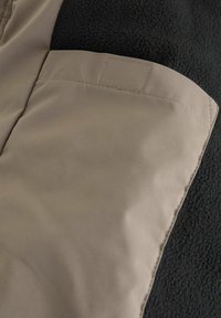 Poche en tissu beige sur une veste en polaire noire texturée ; présente des bords lisses et des coutures visibles, avec un rabat pour un rangement sécurisé.