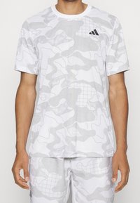 Mężczyzna w białej i jasnoszarej koszulce sportowej z krótkim rękawem w kamuflażowy wzór oraz pasujących szortach, z małym czarnym logo Adidas na klatce piersiowej.