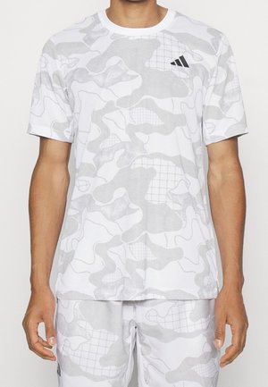 Hombre vestido con una camiseta deportiva de manga corta de patrón de camuflaje blanco y gris claro, y pantalones cortos a juego, con un pequeño logo negro de Adidas en el pecho.