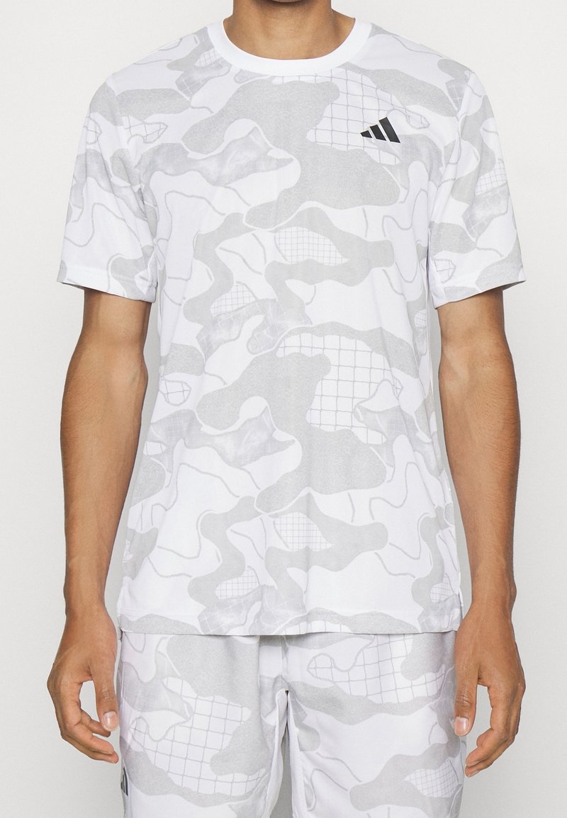 Mężczyzna w białej i jasnoszarej koszulce sportowej z krótkim rękawem w kamuflażowy wzór oraz pasujących szortach, z małym czarnym logo Adidas na klatce piersiowej.