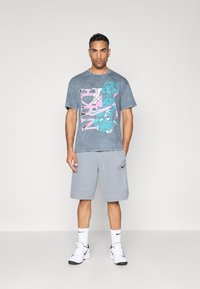 Siva majica s tie-dye uzorkom s ružičastim i plavim grafičkim logotipom, uparena s svijetlosivim šortsevima, bijelim čarapama i bijelim tenisicama s crnim akcentima.