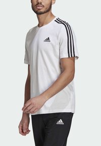 Witte katoenen t-shirt met zwart Adidas-logo en drie zwarte strepen op de schouders. Model draagt ook zwarte broek met Adidas-logo.