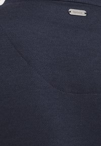 Námořnická modrá tkanina s texturovaným povrchem, obsahující stříbrnou značku Barbour. Jednoduchý, decentní design bez viditelných vzorů nebo dekorací.