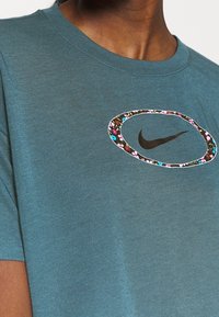 T-shirt in cotone turchese con il logo Nike nero al centro, circondato da un bordo a motivo floreale. Design a maniche corte e collo rotondo.