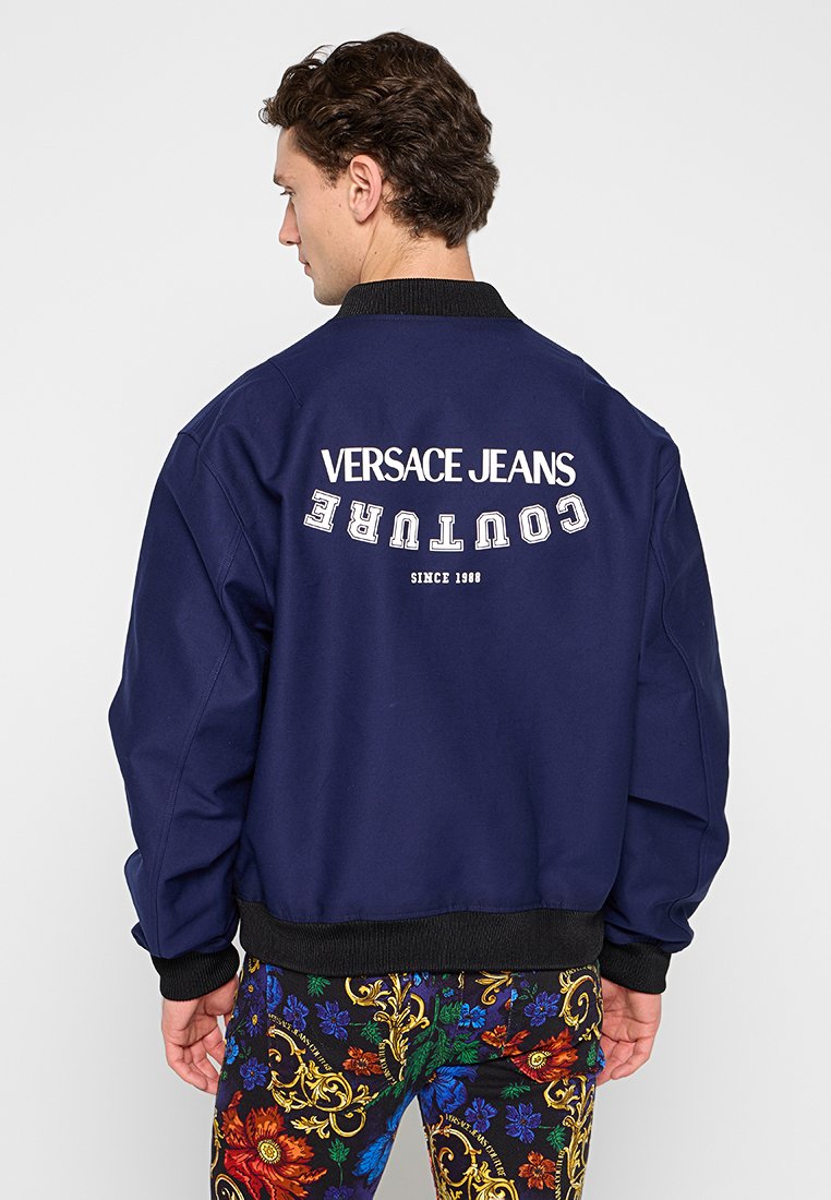Marineblaue Bomberjacke mit geripptem Kragen und Bündchen; auf der Rückseite befindet sich das Logo "Versace Jeans Couture" in weißer Schrift.