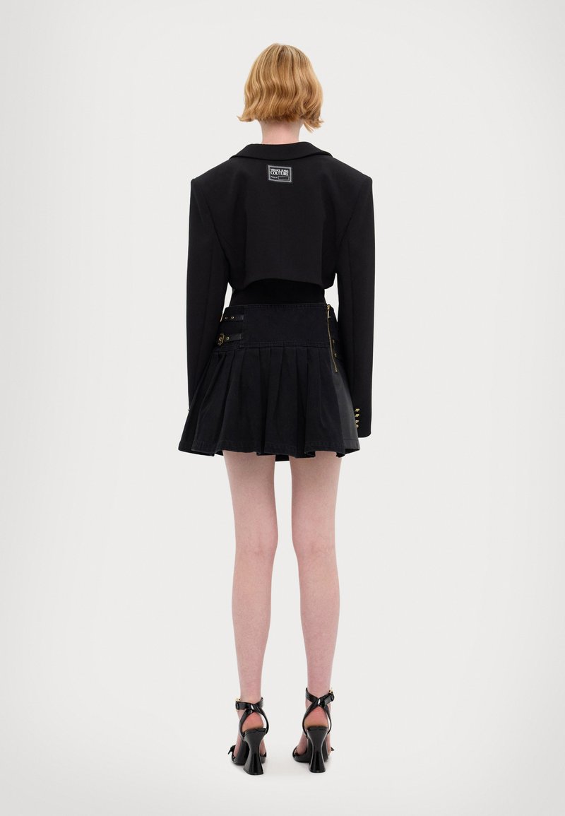 Blazer nero accorciato con spalle ampie, abbinato a una gonna plissettata nera con dettagli in zip e cinghie. Sandali neri con tacco alto.