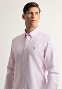 Camisa de botones de rayas color rosa claro, con cuello, mangas largas y pequeño logo en el pecho. Presenta botones en la parte frontal y en los puños.