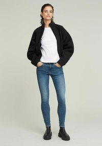 Fekete zippzáras bomber dzseki, oversize ujjakkal, fehér pólóval és kék skinny farmerral párosítva. Fekete vastag bokacsizma zárja le a megjelenést.