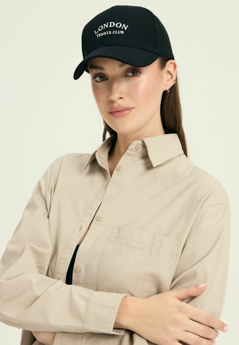 Czarna czapka bejsbolowa z haftem "LONDON TENNIS CLUB" w białym kolorze; noszona z jasnobeżową koszulą z guzikami z kieszonką na piersi.