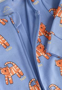 Tutina azzurro chiaro con zip, caratterizzata da motivi di tigre arancione con accenti neri. Tessuto morbido con polsini a coste e zip integrata.