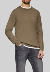 Olive green sweater with a crew neckline, ribbed cuffs, and hem. Worn over a white shirt, paired with faded gray jeans.