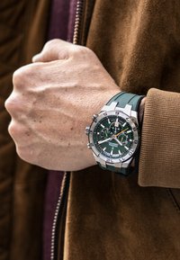 Zaļš un sudraba sporta pulkstenis ar teksturētu gumijas siksniņu, lieliem ciparnīcām un gaismas marķējumiem uz rokas, ko valkā kopā ar brūnu žaketi.