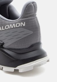 Chaussure de sport grise avec une tige texturée, un col rembourré et une semelle noire robuste. Présente un logo distinct et des éléments de design élégants et angulaires.
