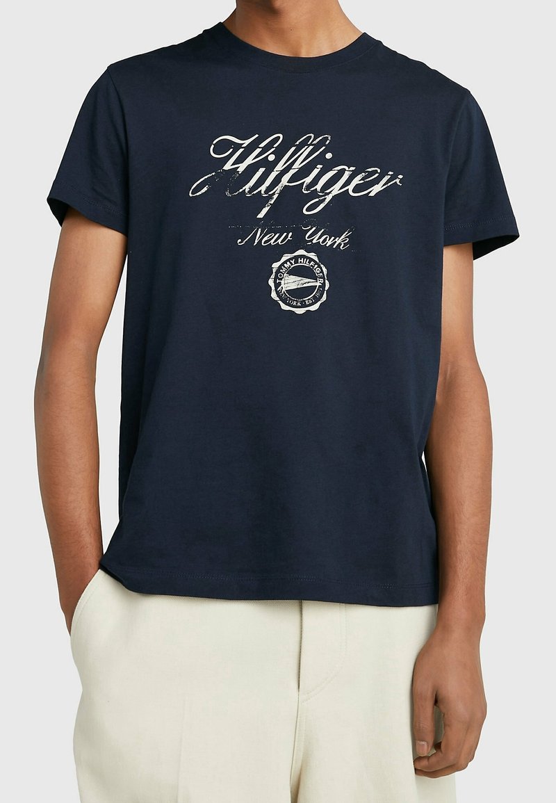 Marinblå bomullst-shirt med en vit logotypdesign som inkluderar "Hilfiger New York" och en cirkulär emblem i mitten. Kortärmad.