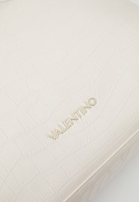 Krämfärgad faux läderhandväska med krokodilstruktur, präglad logotyp i guld och en strukturerad design.