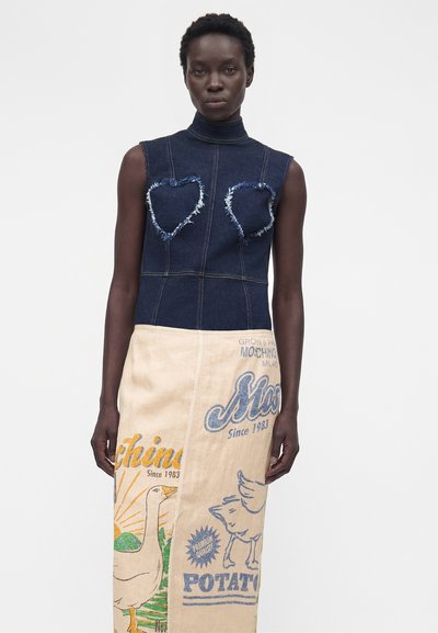 Γυναίκα που φοράει αμάνικο σκούρο ντένιμ τοπ με σκισμένες καρδιές και μπεζ φούστα με πολύχρωμο τυπωμένο κείμενο και εικονογραφήσεις πουλιών.