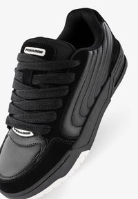 Black athletic shoe featuring a combination of smooth leather and suede materials, thick black laces, and a white rubber sole with textured accents.
