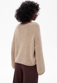 Beżowy sweter z dzianiny o długich, szerokich rękawach i luźnym kroju. Teksturowany materiał, prążkowany dekolt i lekko skrócony tył. W połączeniu z brązowymi spodniami.
