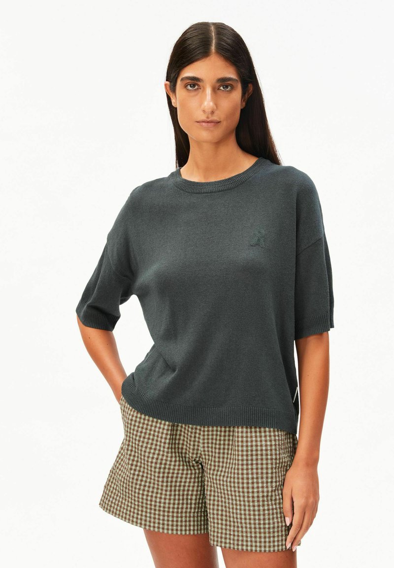 Jonge vrouw met lang donker haar, gekleed in een donkergrijze, korte mouwen gebreide top en bruine geruite korte broek, staand met één hand in haar zak.