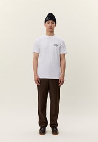 Vīrietis, kurš valkā melnu cepuri, balti T-kreklu ar mazu uzrakstu krūtīs, brūnas bikses un tumšas kurpes, stāv pret vienkāršu gaišu fonu.