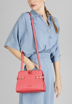 Rosa Lederhandtasche mit goldenen Beschlägen, strukturiertem Design, verstellbarem Tragegurt und Logodetail auf der Vorderseite, gehalten von einer Person.