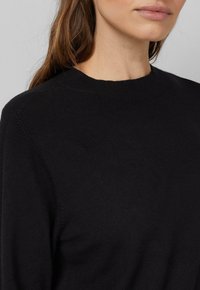 Frau trägt einen schwarzen Rundhalsausschnitt-Pullover, der den Ausschnitt, die Schulter und einen Teil ihres Gesichts sowie braunes Haar zeigt.