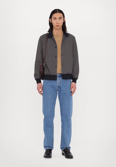 Υφαντό σακάκι σε μαύρο και γκρι χρώμα με γιακά, συνδυασμένο με καφέ μπλουζάκι, μπλε τζιν και μαύρα παπούτσια, κρατώντας μία καφέ τσάντα ώμου.