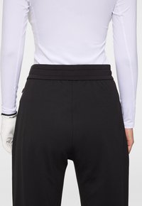 Person som bär svarta byxor, vit långärmad skjorta och vit golfhandske, sedd från midjan till halva låret bakifrån.