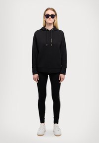 Crna kaputirana hoodie s unutarnjom vrpcom, uparena s crnim tajicama i bijelim tenisicama. Model nosi velike crne sunčane naočale.