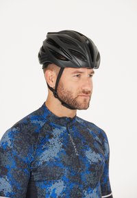 Sort cykelhjelm med ventilationskanaler. Båret af en mand i en blå og sort mønstret cykeltrøje. Højkontrastdesign, aerodynamisk form.