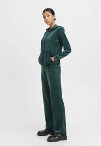 Groene velourse trainingsset met een ritsloze hoodie, zijzakken en bijpassende broek, met een zachte textuur en een wijd uitlopende snit.