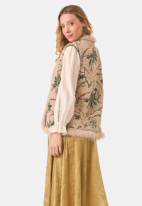 Chaleco beige con bordado floral verde y ribete de piel suave, combinado con una blusa blanca y una falda dorada, mostrando capas de texturas.