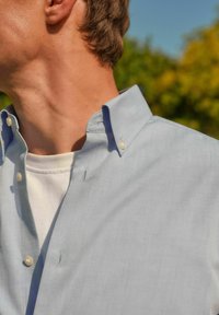 Helles, blaues Hemd mit klassischem Kragen, ausgestattet mit kleinen weißen Knöpfen und einer subtilen Textur, über einem weißen T-Shirt getragen.