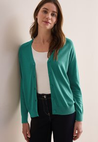 Turkooise, knoopsluiting cardigan in zachte brei stof met lange mouwen en een geribbelde zoom. Draag over een wit shirt en donkere jeans.