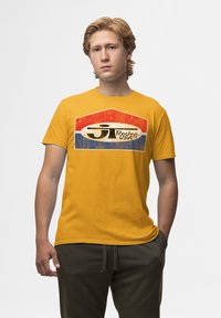 Žluté bavlněné tričko s poškozeným závodním logem v červené, bílé a modré barvě na přední straně. Krátké rukávy a klasický kulatý výstřih.