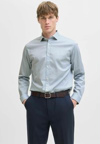 Camicia button-up a quadretti blu chiaro con una texture morbida, maniche lunghe e colletto classico, abbinata a pantaloni scuri e una cintura marrone.